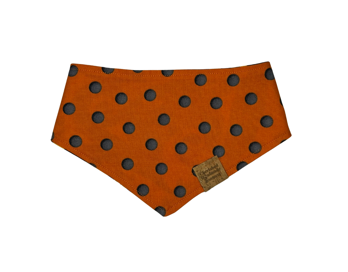 An orange fabric pet bandana with black polka dots, featuring a small brown leather brand tag that reads 'Cherished Handmade Treasures'.