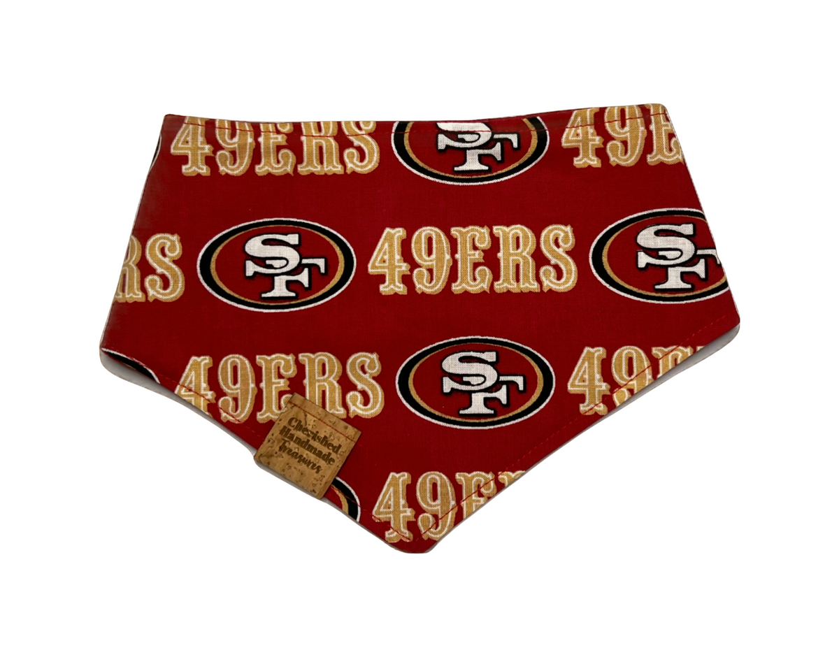 A red pet bandana featuring the San Francisco 49ers logo pattern, with a small brown fabric tag attached near the bottom left corner, shown on a plain white background.
