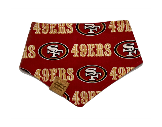 A red pet bandana featuring the San Francisco 49ers logo pattern, with a small brown fabric tag attached near the bottom left corner, shown on a plain white background.